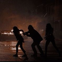 three people standing in the street at night with their arms out and one person holding an umbrella