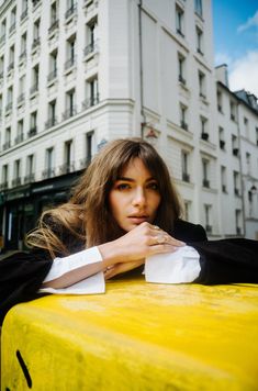 a woman leaning on the edge of a yellow car in front of a tall building