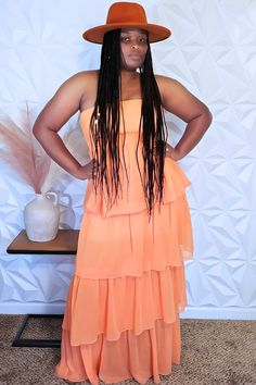 A person confidently stands with hands on hips, wearing the Cabo Rojo Boho Dress from I Am Lilou Bloom. The dress features an orange strapless design with tiered ruffles, complemented by an orange hat. Their long braided hair flows gracefully as they pose in front of a geometric white wall, next to a small table holding a white vase filled with pampas grass. Flowy Tiered Summer Dress, Beach Tiered Dress With Ruffled Skirt, Chic Strapless Sundress With Ruffles, Summer Strapless Tiered Dress With Ruffle Hem, Summer Strapless Tiered Skirt Dress With Ruffle Hem, Strapless Sundress With Smocked Bodice For Brunch, Strapless Tiered Skirt Dress With Ruffle Hem For Summer, Smocked Bodice Tiered Dress For Vacation, Tiered Dress With Smocked Bodice For Vacation