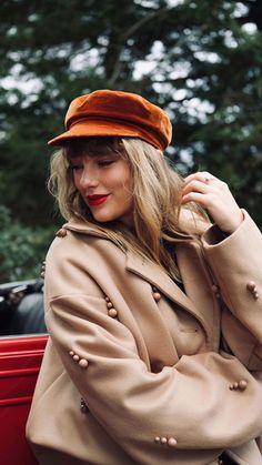 a woman wearing a tan coat and orange hat sitting on a red car with trees in the background