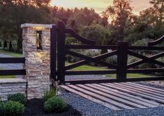 a wooden gate with stone pillars leading to a grassy area at sunset or dawn in the background