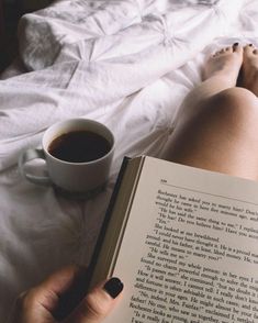 a person laying in bed reading a book and holding a cup of coffee