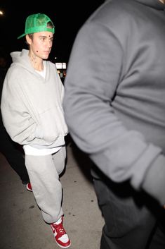 a young man wearing a green hat and grey sweatpants is walking down the street