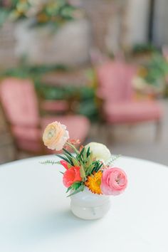 an arrangement of flowers in a white vase on a table with pink chairs behind it