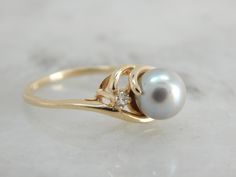 Simple and classic, this pearl ring has a well proportioned gold band, and four balanced prongs set on each side of the pearl. The pearl has a creamy grey luster with pink undertones and makes the perfect understated piece. Metal: 14K Yellow Gold Gem: Grey Pearl Gem Measurements: 6.4 mm, Round Accents: Diamond .04 Carat Ring Size: 4.25 Marks: "14K" Stamped on the inside band Luxury Heirloom Pearl Ring With Prong Setting, Pearl Grey, Lovely Jewellery, The Pearl, Hand Engraving, Pearl Ring, Gold Band, Cocktail Ring, A Well