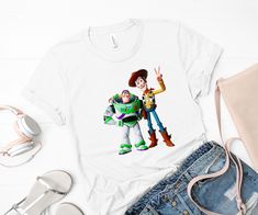 "This Toy Story tee is a super-soft, baby-knit t-shirt looks great on both men and women. It is an updated unisex tee that's printed with high quality ink, which fits like a well-loved favorite. Made from 100% cotton, the double-stitched neckline and sleeves give it more durability. It's part of the Bella and Canvas line, so it's softer and more fitted than other tee styles. It's also preshrunk, but can size down a half size if dryed in high heat. SIZING GUIDE (chest width AROUND in inches - Measure around chest at largest part) *Shirts are UNISEX sizing.* S: 36 M: 40 L: 44 XL: 48 2XL: 52 3XL: 56 PRODUCT CARE Feel free to wash, dry or hang any of our products as you normally would the rest of your wardrobe! Specific care instructions are printed by the manufacturer onto the tag however! SP Casual T-shirt With Character Print For Playtime, Themed Short Sleeve Tops For Playtime, Playful Tops With Character Print For Fan Merchandise, White Novelty Tops With Character Print, Themed White Tops With Character Print, Playful Cartoon Print Top For Fans, Trendy Graphic Print T-shirt For Playtime, Toy Story Shirt, Woody And Buzz