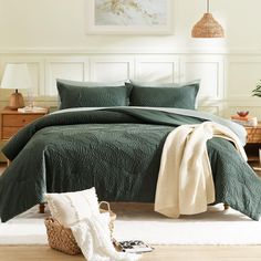 a bed with green bedspread and pillows in a white room next to a wicker basket