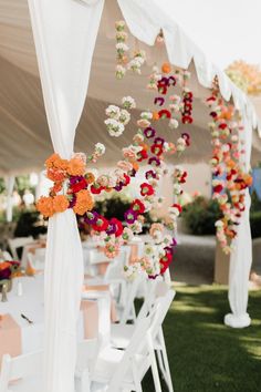 an outdoor wedding with white chairs and flowers on the back wall, along with orange and pink blooms