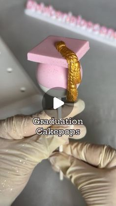 a person in gloves is holding a pink and gold graduation cakepops with a golden ring on it