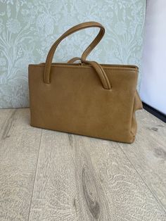 This is a stylish ladies handbag in a camel or beige leather-look material that has a smooth texture. It has a rectangular shape and a vintage look that evokes the elegance of the mid-century. The inside is lined with black satin that feels soft and luxurious. It has two side zipped pockets that are spacious and secure, and a middle section with a small zipped pocket that is perfect for storing small items. The frame, clasp and feet have gold accents that contrast beautifully with the neutral collar of the bag. The size is 28 cm wide, 16.5 cm high and 9 cm deep at the base, making it a versatile accessory that can fit many essentials.  The frame has the mark of Elbief England, a company that made frames for high quality British handbags from the 1940s to the 1970s. The mark is a sign of cr Classic Beige Bags With Hasp Closure, Classic Beige Bag With Hasp Closure, Light Brown Rectangular Satchel For Shopping, Chic Camel Shoulder Bag For Evening, Classic Beige Box Bag With Handles, Beige Square Satchel With Hasp Closure, Light Brown Rectangular Satchel With Detachable Handle, Beige Evening Box Bag With Handles, Elegant Camel Top Handle Satchel