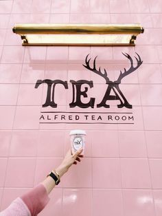 a person holding up a cup in front of a pink wall with the word tea on it