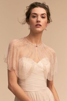 a woman wearing a white dress with sheer sleeves and beading on the shoulders, in front of a beige background