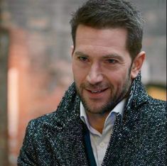 a man wearing a jacket and tie standing in front of a brick building smiling at the camera