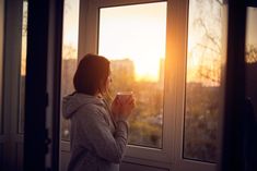 a woman standing in front of a window holding a coffee cup and looking out the window