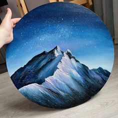 a person holding up a painting of mountains and stars in the night sky with their hand