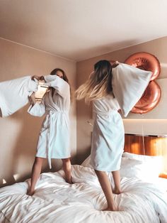 two women in bathrobes standing on a bed with white sheets and brown pillows