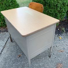 an old desk with a wooden chair next to it on the sidewalk in front of some bushes