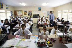 a classroom full of students sitting at desks with their teacher standing in the background