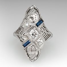 This striking circa 1930s dinner ring features a pierced design with engraved details and bordered with milgrain edging. The ring is accented with three (3), bead set, round transitional brilliant cut diamonds and two (2), bezel set, rectangular French cut synthetic sapphires. The ring measures 23.2mm at the top, rises 5.8mm above the finger, tapering to 1.4mm wide and 0.8mm thick at the base of the shank. This ring is currently a size 3 and can be sized larger. Art Deco Three Stone Platinum Jewelry, Elegant Three Stone Collectible Ring, Vintage Three-stone Platinum Jewelry, Vintage Three Stone Platinum Jewelry, Art Deco Three Stone Ring For Formal Occasions, Vintage Platinum Three-stone Jewelry, Formal Art Deco Three-stone Diamond Ring, Art Deco Diamond Cut Jewelry For Vintage Events, Art Deco 14k White Gold Jewelry