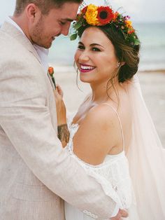 a bride and groom standing on the beach with their arms around each other, smiling