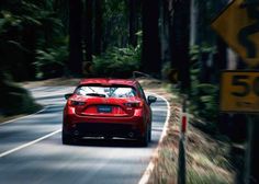 a red car driving down a road next to a forest filled with lots of trees