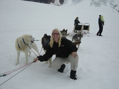 a woman kneeling down next to two dogs on a snow covered slope with people in the background