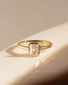 a gold ring with an emerald cut diamond on the front and side, sitting on a white surface