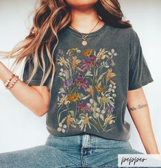 a woman with long hair sitting on the ground wearing a t - shirt that has flowers painted on it