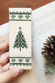 a hand holding a bookmark with a cross stitch christmas tree on it and pine cones next to it