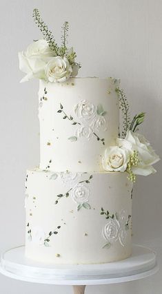 two tiered wedding cake with white flowers and greenery on the top, sitting on a stand