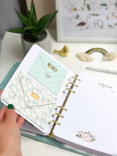 a person holding an open planner book on top of a desk next to a potted plant