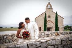 a bride and groom standing in front of a stone wall with a church behind them