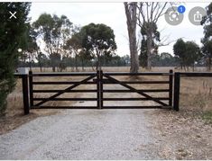 a wooden gate in the middle of a dirt road with trees and grass behind it