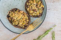 two stuffed mushrooms are on a blue plate with a gold fork and sprig of rosemary