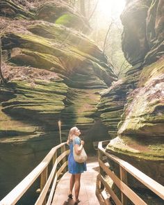 a woman standing on a wooden bridge in the middle of a forest with large rocks behind her