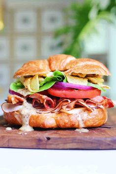 a sandwich with meat, lettuce and tomato on a wooden cutting board next to a potted plant