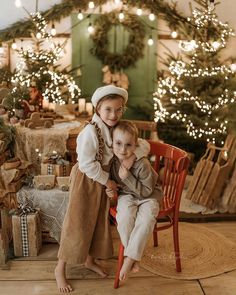 two children sitting on a chair in front of christmas trees