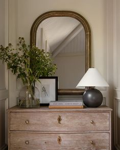 a wooden dresser topped with a lamp next to a mirror and vase filled with flowers