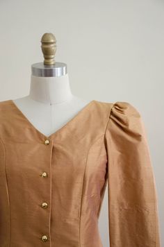 A homemade golden tan jacket, with puffed shoulders, gold faceted buttons, and good tailoring. It is unlined and light enough to be worn as a blouse. ☛ m e a s u r e m e n t s ☚ Bust: 32 Waist: 28 Shoulders: 13.5 Length: 21 ☛ d e t a i l s ☚ Era: 1990s Material: polyester Condition: excellent ☛ v i s i t t h e s h o p ☚ https://etsy.me/2Nd23kg ☛ instagram ┇ poppycockvintage ☛ facebook ┇ poppycockvintage Fitted Tops With Gold Buttons For Workwear, Fitted Gold Top For Workwear, Spring Gold Blouse With Buttons, Fitted Gold Tops For Work, Fitted Vintage Gold Blouse, Vintage Gold Blouse For Spring, Spring Vintage Gold Blouse, Vintage Gold Fitted Blouse, Fitted Gold Vintage Blouse