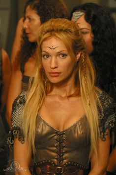 a woman with long blonde hair wearing a leather dress and chain around her neck, standing in front of other women