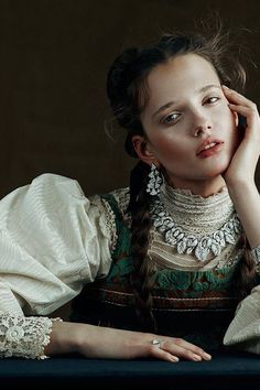 a woman with braids sitting at a table wearing a white dress and holding her hand to her face