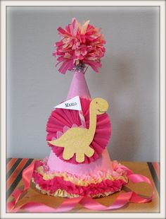 a pink and gold birthday hat with a yellow dinosaur on it's side, sitting on top of a table