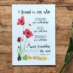 a card with flowers on it sitting next to a green plant and a piece of paper that says, a friend is one who