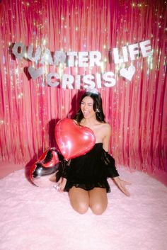 a woman sitting in front of a pink backdrop holding two heart shaped balloons