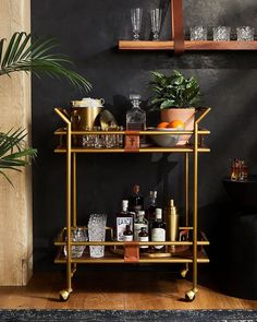 a gold bar cart with drinks on it in front of a black wall and potted plant