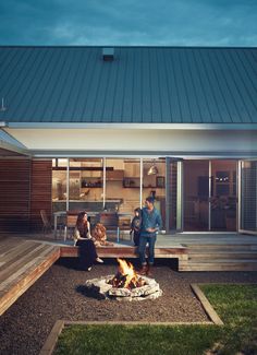 three people sitting around a fire pit in front of a house with sliding glass doors