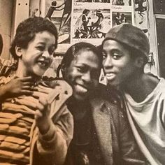 black and white photograph of three young men posing for the camera in front of a wall with posters on it