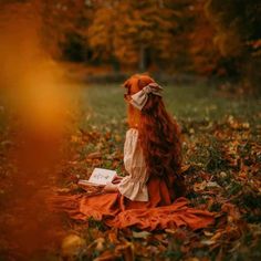 a woman sitting on top of a field covered in leaves reading a book next to trees