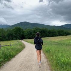 a woman running down a dirt road in the middle of a field with mountains in the background