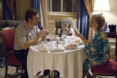 a man and woman sitting at a table eating food with a dog in the background